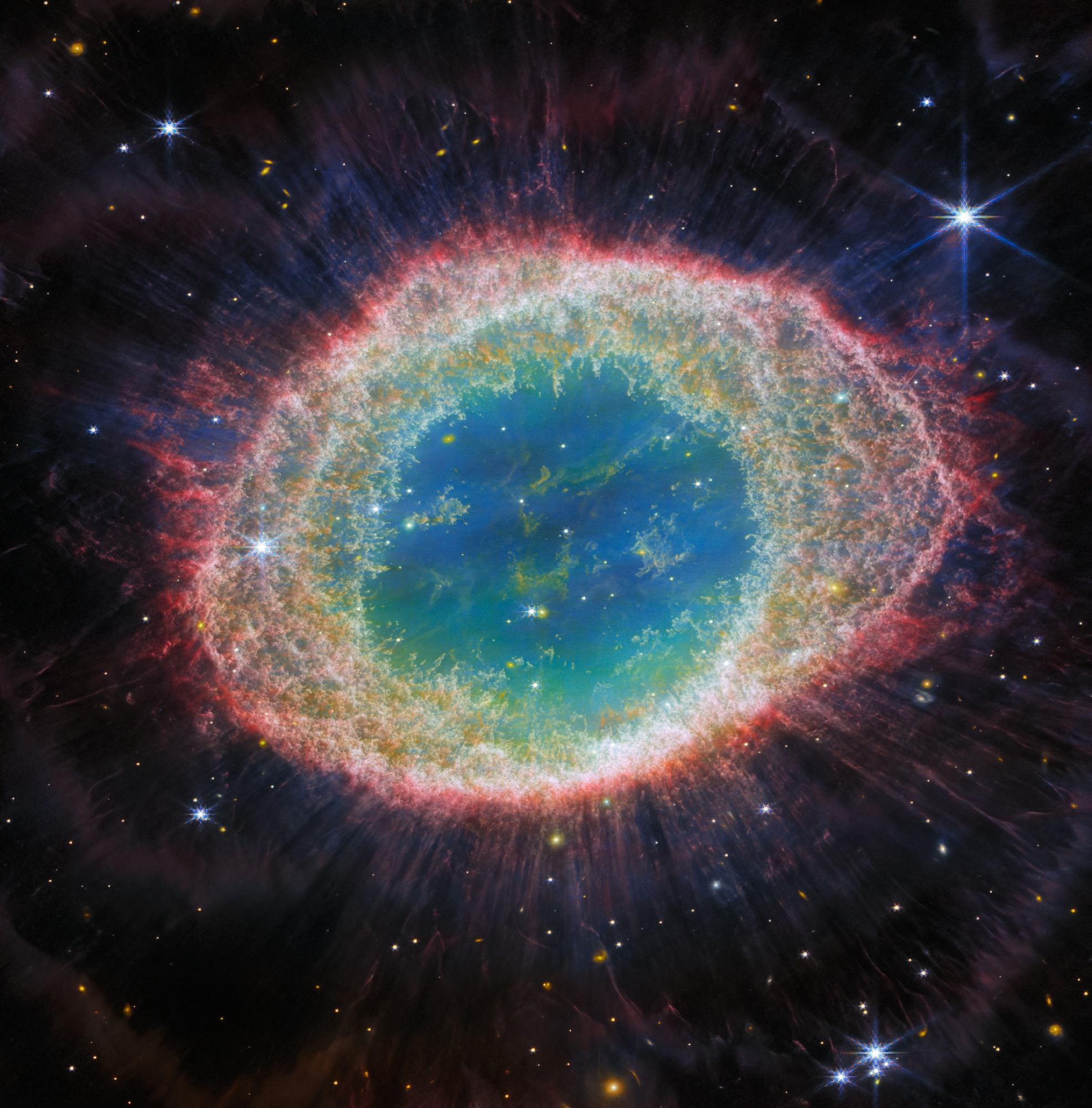 Na ilustracji: Zdjęcie ikonicznej Mgławicy Pierścień (M57, NGC 6720) wykonane przez Teleskop Webba współpracujący z kamerą NIRCam (Near-Infrared Camera) o bezprecedensowej szczegółowości. Mgławica Pierścień jest archetypem mgławicy planetarnej, która powstała, gdy zaawansowana wiekiem gwiazda odrzuciła otoczkę z powodu braku paliwa jądrowego. Na tym zdjęciu  w bliskiej podczerwieni szczególnie dobrze widać misterną strukturę włókien wewnętrznego pierścienia. Widać około 20 tysięcy gęstych globuli bogatych w molekularny wodór. W przeciwieństwie do tego, w centralnym obszarze znajduje się bardzo gorący gaz. Główna otoczka  jest delikatnym pierścieniem, w którym intensywnie świecą molekuły zawierające węgiel – takie jak np. wielopierścieniowe węglowodory aromatyczne PAH. Źródło: ESA/Webb, NASA, CSA, M. Barlow (University College London), N. Cox (ACRI-ST), R. Wesson (Cardiff University